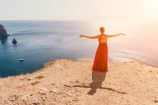 Side view a Young beautiful sensual woman in a red long dress posing on a rock high above the sea during sunrise. Girl on the nature on blue sky background. Fashion photo.