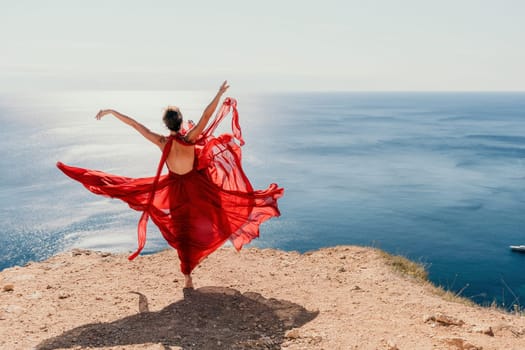 Side view a Young beautiful sensual woman in a red long dress posing on a rock high above the sea during sunrise. Girl on the nature on blue sky background. Fashion photo.