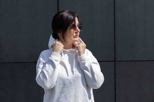 Portrait Mature Brunette Woman With Hood On Head, Wears White Hoody and Sunglasses, Dark Granite Background Wall. Sportive Confident 40 Yo Beautiful Caucasian Woman Outdoor. Horizontal Plane.