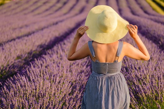Standing amidst the vibrant lavender field, a woman wearing a sundress gently places her hands on her straw hat, basking in the beauty of nature on a sunny summer day, backside