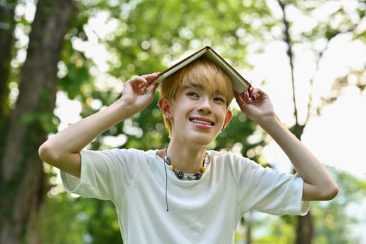 University student man putting open book on head, reading book in public park on sunny day.