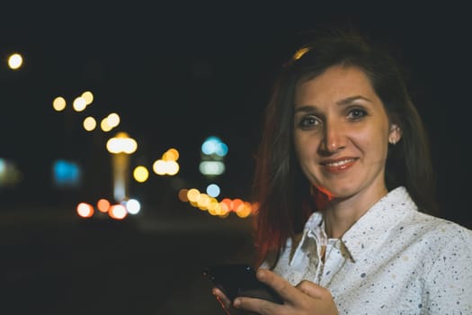 attractive girl in white suit with smartphone in night light reflection standing near copy space area for advertising, young woman smiling with mobile in hands