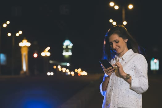 attractive girl in white suit using smartphone in night, light reflection with copy space area for advertising, young woman smiling with mobile in hands