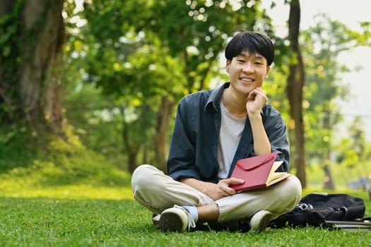 Smiling asian student man sitting on the grass in the city park and reading a book. Youth lifestyle and education.