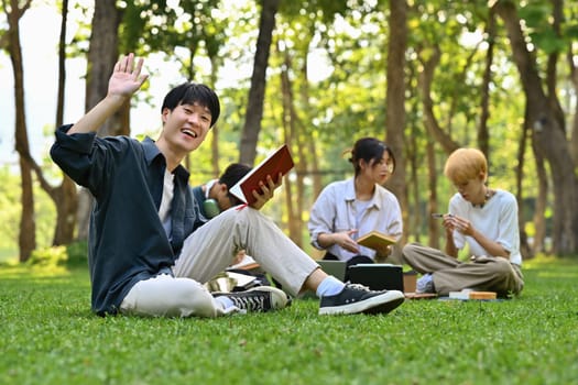 Smiling asian man student sitting on green grass in campus with friends on background. Education, technology and lifestyle.