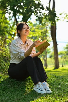 Pleasant young woman reading favorite book, sitting on a green meadow in beautiful summer day. Education, learning and lifestyle.