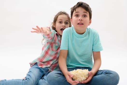 Adorable two diverse children with bowl of popcorn, looking toward the camera with frightened look, watching scary or fantasy movie, expressing fright and fear, isolated over white studio background