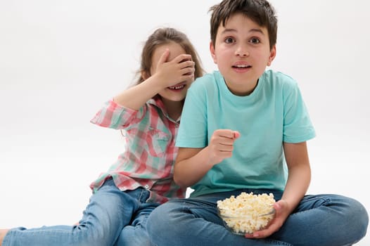 Amazed kids, teen boy and little child girl covering her eyes from fright, watching scary movie or cartoon, eating popcorn over white isolated background. People. Kids. Leisure activity. Entertainment