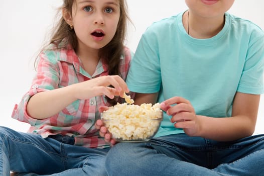 Cropped view of adorable diverse kids with a bowl of popcorn, looking fascinated surprised astonished while watching movie or cartoon, isolated on white studio background. People. Leisure. Lifestyle