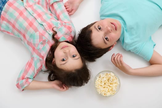View from above of Caucasian adorable kids, beautiful little girl in checkered shirt and handsome preteen boy in blue t-shirt, lying on white background with a bowl of popcorn, looking at camera