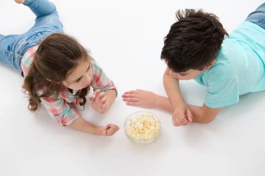 Top view of lovely children - preteen boy and cute baby girl, brother and sister eating popcorn, lying on white isolated studio background. Kids. People. Lifestyle. Leisure activity. Entertainment