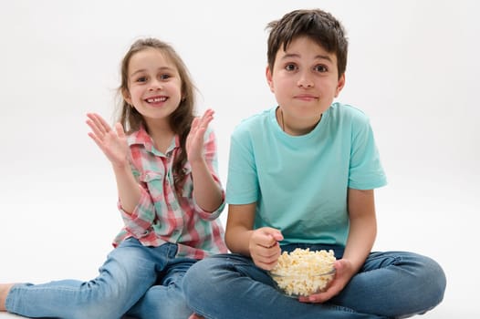 Portrait on white background of beautiful kids, teenager boy and preschooler girl, older brother and younger sister eating popcorn, expressing happy positive emotions, isolated on white background