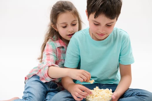 Lovely children - preteen boy and cute baby girl, brother and sister eating popcorn, isolated on white studio background. Kids. People. Lifestyle. Leisure activity. Entertainment. Family relationships