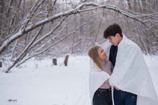 A young couple walks in the park in winter. The guy and the girl are kissing wrapped in a white blanket outdoors
