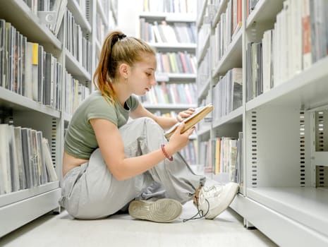 Teenager girl sitting in library on floor and reading academic book. Beautiful female student preparing for exam with education mterials in bookstore