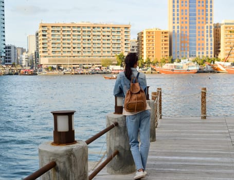 A young woman with a backpack on her back admires the river in the old Dubai Creek. Back view. Journey through the Persian Gulf.