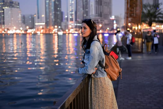 A tourist girl with a backpack on her shoulders enjoys a view of the modern skyscrapers of the Sharjah marina at night.