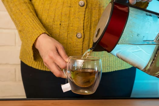 Close-up of a woman pouring hot water from a kettle into a clear glass mug. a cup of freshly brewed herbal tea. The process of brewing tea or tea ceremony in a warm soft light. Movement of boiling water pouring into a cup