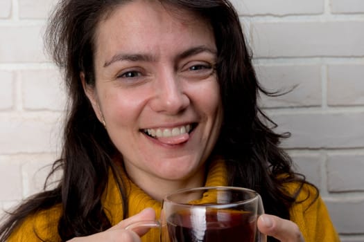 a pretty Caucasian middle - aged woman drinks tea and laughs playfully showing her tongue while spending time with friends . transparent mug with delicious fragrant tea. Enjoying the moment, mental health and positive emotions