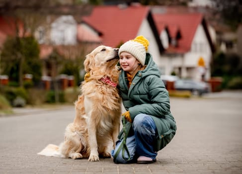 Preteen child girl sitting with golden retriever dog at autumn city street wearing hat and warm jacket.
