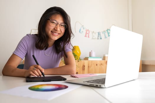 Asian young female graphic design student working on a project at home using tablet and laptop looking at camera. Color wheel. Creative occupation.