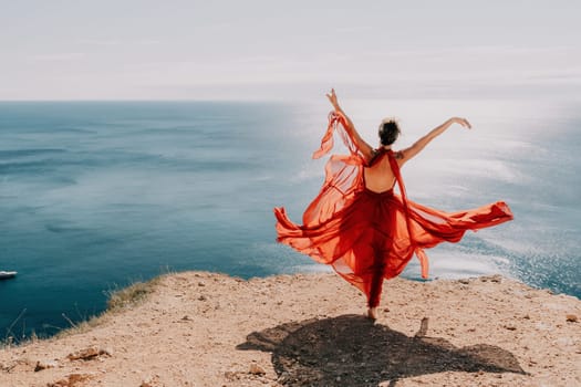 Side view a Young beautiful sensual woman in a red long dress posing on a rock high above the sea during sunrise. Girl on the nature on blue sky background. Fashion photo.