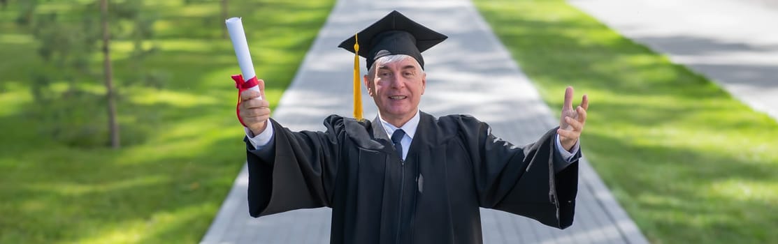 An elderly gray-haired man in a graduate robe spread his arms to the sides and holds a diploma outdoors. Widescreen