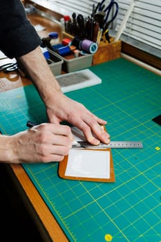 A man's hand holds pieces of leather for a leather wallet in his workshop. The process of working with natural brown leather. The craftsman holds the craft