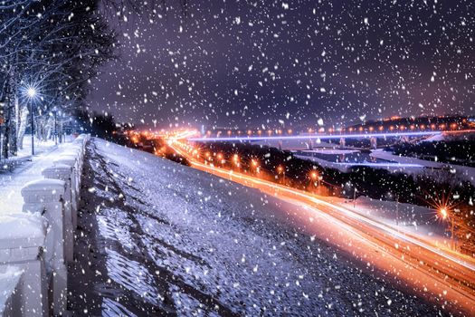 Snowfall in a winter park at night with lanterns, view to road with car motion, pavement and trees covered with snow.