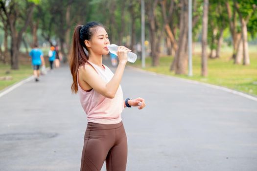 Asian woman with pink sportswear stand on road in park and drink water from bottle during exercise in the morning.