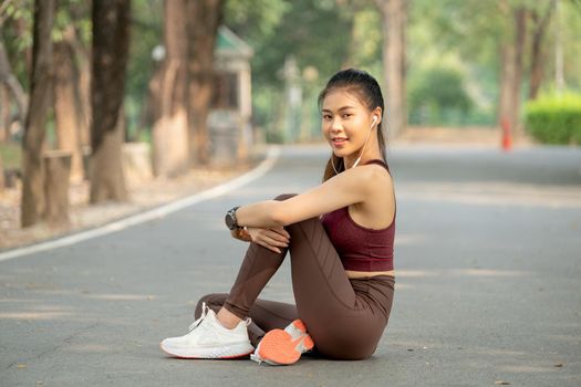 Asian sport woman sit and hug the knee also look at camera with smiling during exercise in park or garden in the morning.