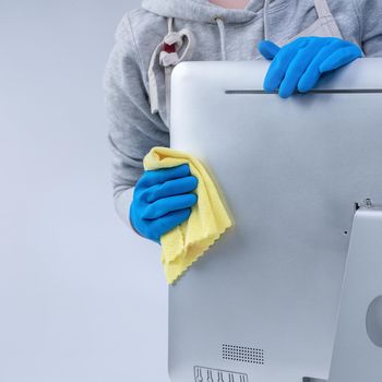 Young woman housekeeper in apron is cleaning silver computer surface with blue gloves, wet yellow rag, close up, copy space, blank design concept.