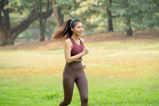 Wide shot of Asian woman jogging in park or garden with green field and tree as background during morning time.