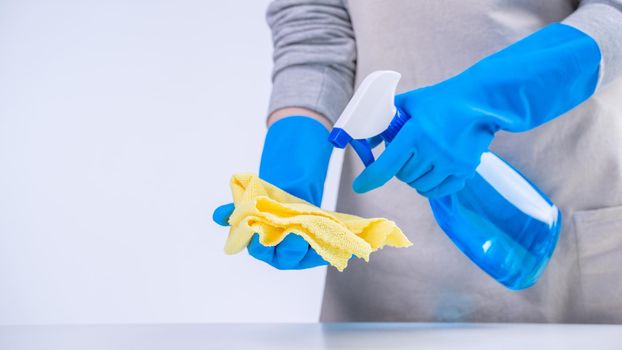 Young woman housekeeper is doing cleaning white table in apron with blue gloves, spray cleaner, wet yellow rag, close up, copy space, blank design concept.