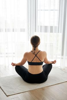 Back View Of Woman Sitting In Yoga Lotus Pose Relaxing And Meditating In Living Room