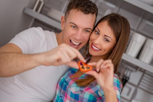 A happy young couple is having fun in the kitchen, holding a slice of red paprika in heart shape. 