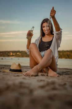 Young woman relaxing at sunset time on the river bank. She is sitting by the river, drinking beer and playing with sand. 