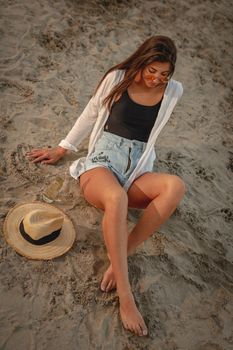 Top view of a young woman with straw hat relaxing at sunset time on the river bank. She is sitting on the send and enjoying. 