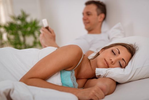 Young woman sleeping while her boyfriend using smartphone in bed. Selective focus.