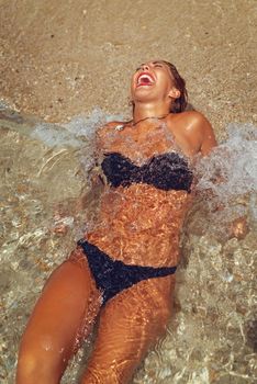 Beautiful young woman enjoying on the beach. She is lounging and sunbathing on the sandy coast in the sea while the waves splash on her body. 