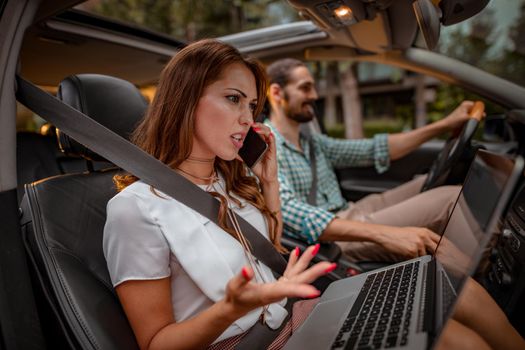 Young confident couple go to business trip, a woman is working on laptop and talking on smartphone, while a man is driving.