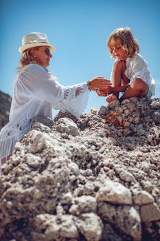 Beautiful little boy enjoying with his mother on the beach rock. The mother gives handful little stones to her son.
