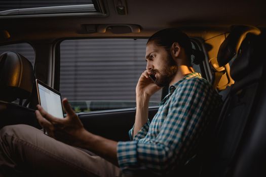 Young confident businessman is holding smartphone, talking and looking at laptop on the back seat in car.