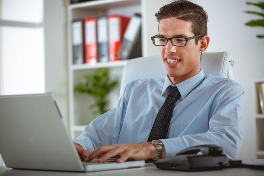 Executive satisfied male working at laptop looking at documents and accounts. He is typing and looking concentrated and serious.