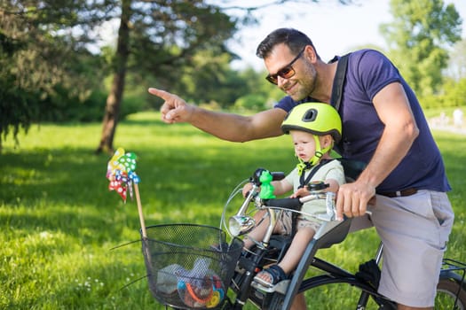 Look over there. Active family day in nature. Father and son ride bike through city park on unny summer day. A cute boy is sitting in front bicycle chair while father rides bicycle. Father son bonding