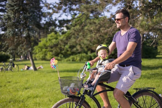 Look over there. Active family day in nature. Father and son ride bike through city park on unny summer day. A cute boy is sitting in front bicycle chair while father rides bicycle. Father son bonding