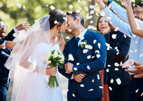 Newlywed couple bride and groom sharing a kiss while surrounded by friends and family and being showered with confetti rose petals after their wedding ceremony.