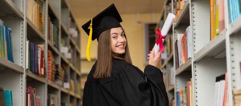 Happy young woman in graduate gown holding diploma in the library. Widescreen