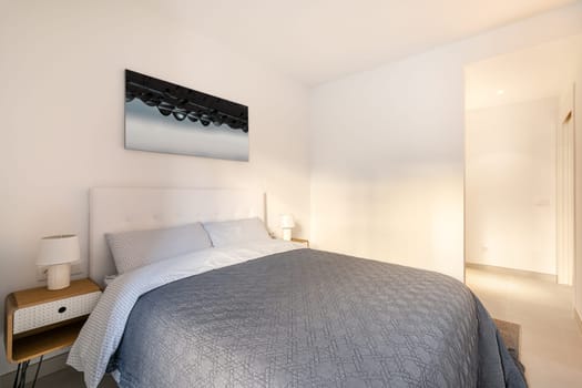 Bedroom with double bed with a picture on the wall and stylish bedside tables in light colors and an open door to the corridor. The concept of a compact hotel room. Copyspace.