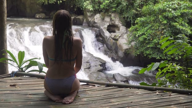 Girl sits on a wooden flooring in rainforest and looks at beautiful waterfall. Bikini girl sitting next to idyllic tropical waterfall. Bali,Indonesia.